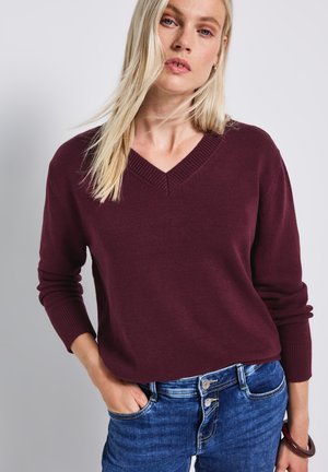 Bordeaux V-hals striktrøje lavet af strikket stof, med ribbede manchetter og kant. Båret med denim jeans, der viser en afslappet pasform.