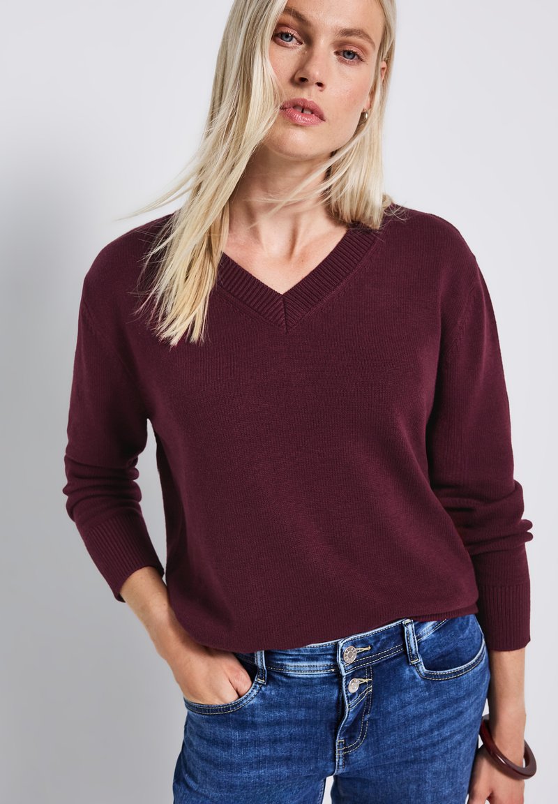 Pull en V bourgogne en tissu tricoté, avec des poignets et un ourlet côtes. Porté avec un jean en denim, montrant une coupe décontractée.
