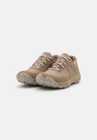 Beige Sportschuhe aus strukturiertem Material mit Schnürsenkeln, grauen Akzenten und einer robusten, gemusterten Laufsohle. An der Seite befindet sich ein kleines oranges Detail.