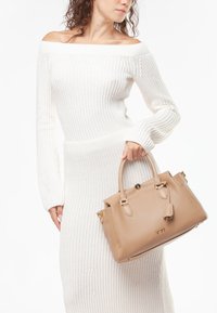 Beige leren handtas met gestructureerde vorm, functionele hardware en een label. Model draagt een witte gebreide off-shoulder jurk.