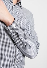 Chemise boutonnée à motifs vichy en bleu marine et blanc. Elle arbore un col et des poignets boutonnés, avec une coupe ajustée et un tissu texturé.