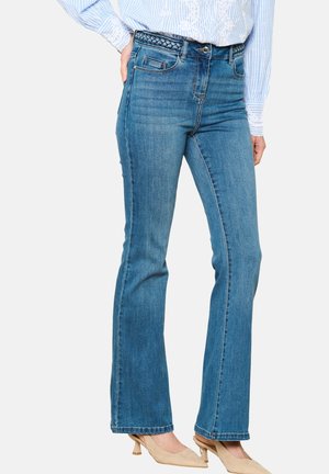 Blauwe denim flare jeans met een middelhoge taille, gevlochten riemdetails en subtiele vervaging. Gecombineerd met beige hakken.