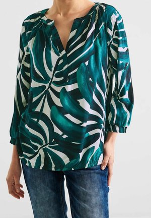 Vrouw die een blouse met lange mouwen draagt met grote groene tropische bladpatronen en een blauwe spijkerbroek tegen een effen achtergrond.