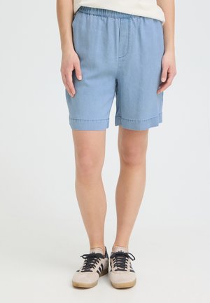 Personne portant un short bleu clair à taille élastique et des baskets beiges avec des rayures noires, debout devant un fond clair uni.