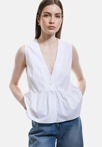 Top sin mangas blanco con escote en V profundo, cierre de botones en el frente, diseño peplum fruncido y ajuste casual, combinado con unos jeans azules holgados.