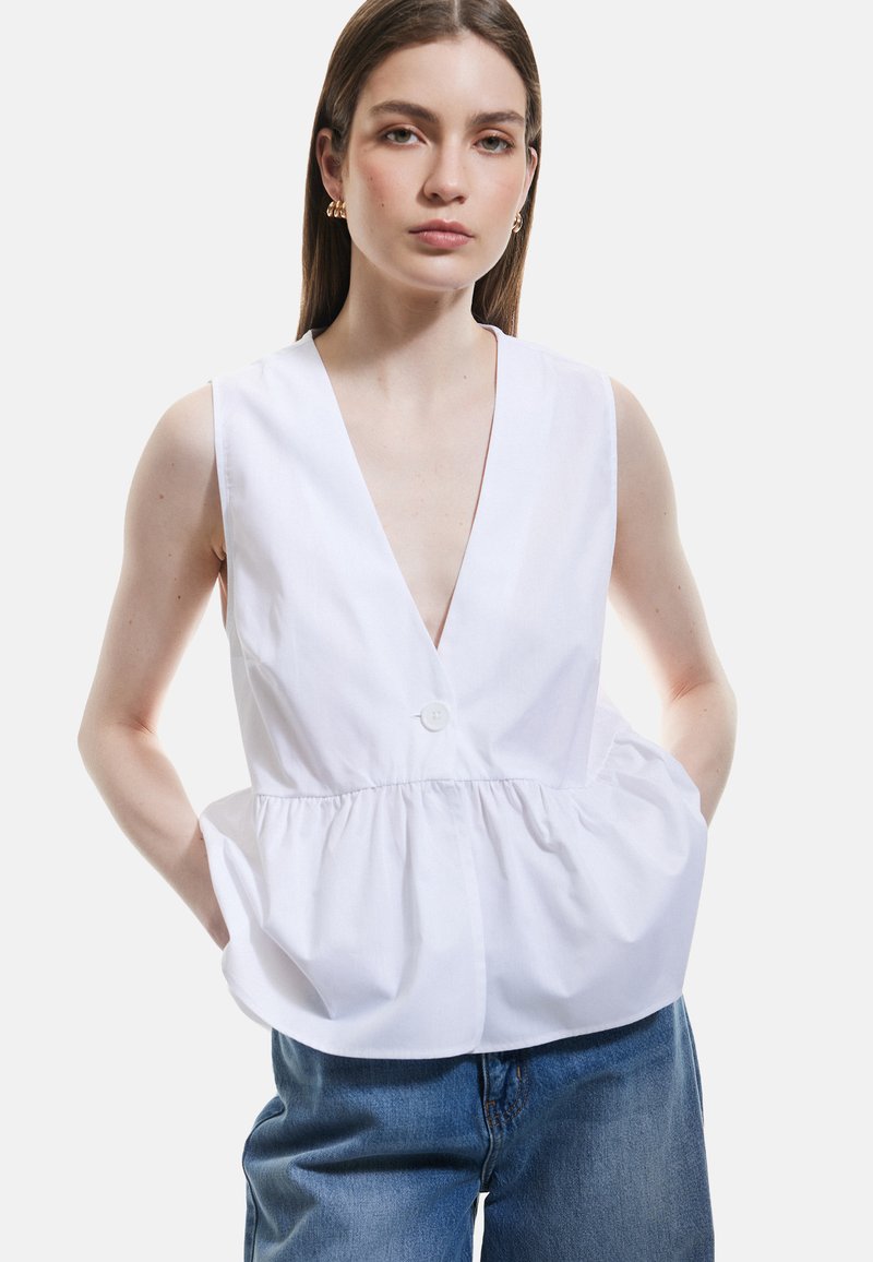 Top sin mangas blanco con escote en V profundo, cierre de botones en el frente, diseño peplum fruncido y ajuste casual, combinado con unos jeans azules holgados.
