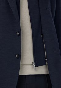 Jūras zilas krēslas ar teksturētu audumu, izceļot melnas pogas un kontrastējošu gaiši bežīgu adītu slāni zemāk, ar redzamu rāvējslēdzēju.