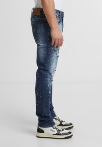 Bleka blå denimjeans med trasiga detaljer, som har stänkmönster, rak passform och midjeband i brunt läder. Matchade med svarta och vita sneakers.