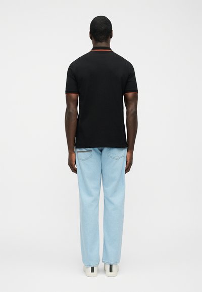 Άνδρας που στέκεται και κοιτάζει προς τα πίσω, φοράει μαύρο κοντομάνικο πουκάμισο με κόκκινη λεπτομέρεια, ανοιχτό μπλε τζιν και άσπρα αθλητικά παπούτσια με μαύρες και πράσινες ρίγες.