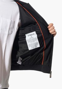 Giacca nera con bordo arancione, fronte con zip, foderata in rete e etichetta di cura visibile all'interno. Presenta una vestibilità slim e polsini elasticizzati.