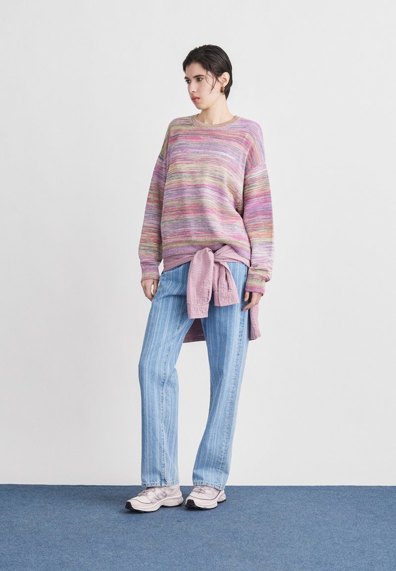 Donna con maglione multicolore pastello, jeans azzurri a righe, camicia rosa legata in vita e sneakers rosa chiaro, in piedi su un tappeto blu.