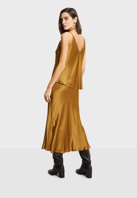 Ensemble en satin de couleur moutarde composé de deux pièces, avec un haut sans manches et une jupe fluide, présentant un dos dénudé et un design superposé, complété par des bottes noires jusqu'aux genoux.