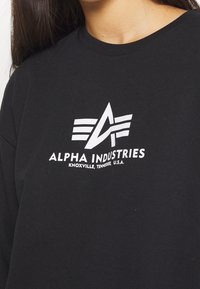 Sweat-shirt noir avec un logo graphique blanc composé de trois bandes stylisées et le texte "ALPHA INDUSTRIES", confectionné en tissu de coton doux.