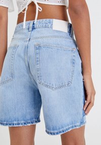 Shorts en denim bleu clair avec ourlets effilochés, poches arrière et un patch en cuir blanc à la taille. Caractéristiques d'une coupe décontractée et d'un tissu texturé.