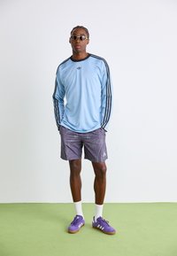 Mies, jolla on päällään vaaleansininen pitkähihaista Adidas-paitaa, purppurakuosiset Adidas-shortsit, purppurat Adidas-urheilukengät, valkoiset sukat ja vaaleat aurinkolasit, seisoo vihreällä lattialla.