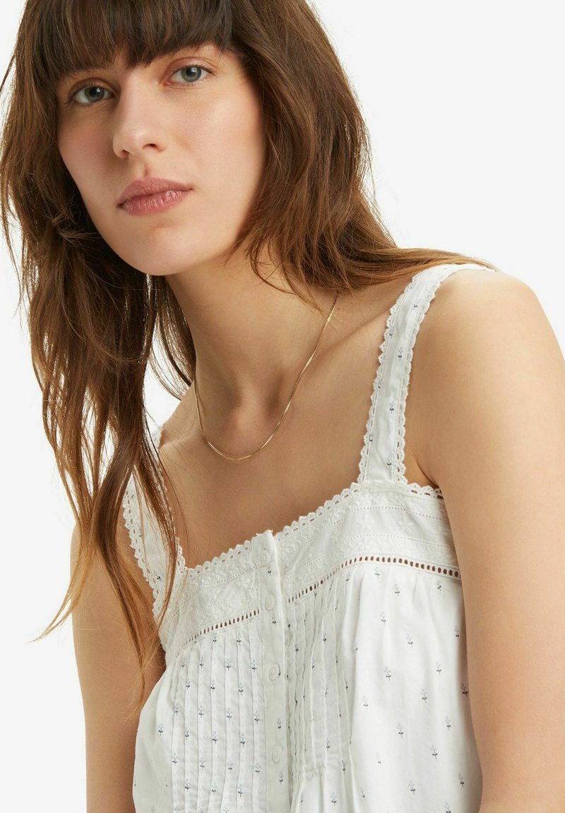 Vestido blanco sin mangas con delicados bordados y un dobladillo de encaje. Presenta botones en la parte delantera y un corte relajado. Collar de cadena dorada sencillo.