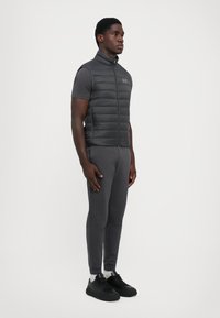 Gilet rembourré gris avec un col montant et une fermeture éclair à l'avant, porté sur une chemise à manches courtes, associé à un pantalon fuselé assorti et des baskets noires.