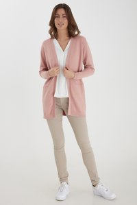 Cardigan rosa chiaro realizzato in morbido materiale a maglia, con apertura frontale, due tasche anteriori e polsini a coste, abbinato a una camicetta bianca e pantaloni beige.