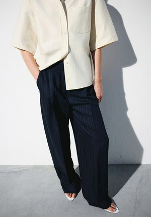 Persona con una camisa cremosa de manga corta y suelta, pantalones anchos a rayas negras, con las manos en los bolsillos, de pie contra una pared blanca.