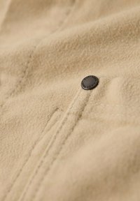 Tan fleece stof met een zachte textuur, voorzien van een zwarte metalen drukknoop met gegraveerde tekst, geplaatst nabij een naad.