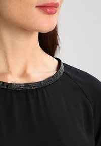 Schwarzes Oberteil mit einem runden Ausschnitt und einem metallischen Perlendekor um den Kragen. Glatter Stoff mit leichtem Glanz.