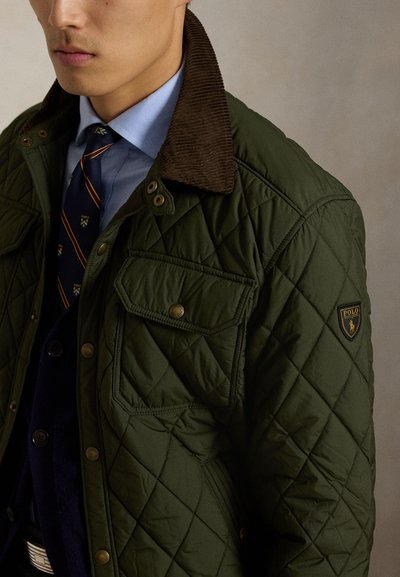 Hombre con chaqueta acolchada verde con cuello de pana marrón sobre camisa azul y corbata a rayas en fondo neutro.