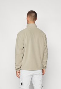 Jachetă din fleece bej cu guler înalt, mâneci lungi și o textură moale, purtată peste pantaloni cargo albi. Vederea din spate arată detalii minime.