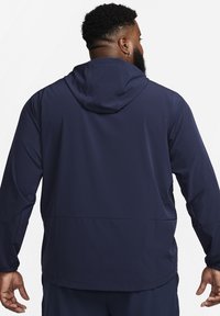 Marinblå huva jacka gjord av mjukt material med elastiska muddar och en bakficka med dragkedja, som visar en enkel och funktionell design.