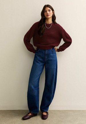 Suéter acanelado burdeos, jeans de pierna ancha de cintura alta en azul, y zapatos planos marrón oscuro. Collares de plata en capas complementan el atuendo.