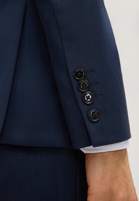Manica di un completo blu navy con tessuto texturizzato, quattro bottoni neri e polsino bianco di contrasto che spunta.
