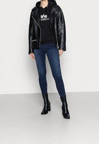 Chaqueta de cuero negra de estilo motero sobre una sudadera con capucha negra, combinada con jeans ajustados oscuros y botines negros de tacón alto. Diseño simple y ajustado.