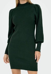 Femme portant une robe en maille côtelée vert foncé avec des manches longues bouffantes et un col montant, debout devant un fond blanc.