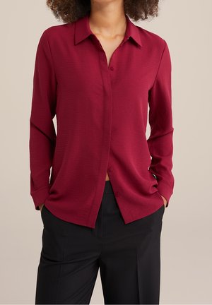 Femme portant un chemisier bordeaux à manches longues boutonné, avec un pantalon noir, les mains dans les poches, sur un fond beige uni.