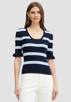 Donna che indossa un top a coste blu navy con righe orizzontali bianche e azzurre e maniche corte arricciate, abbinato a pantaloni bianchi.