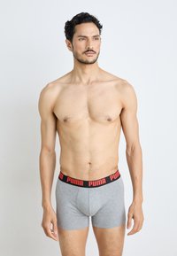 Szürke boxer alsónadrág fekete derékrésszel, piros "PUMA" logóval. A modell egyenesen áll, semleges arckifejezéssel, mutatva a törzsét.