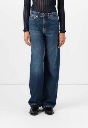 Weite blaue Denim-Jeans mit hoher Taille. Sie verfügen über ein klassisches Fünf-Taschen-Design und eine ausgewaschene Textur. Kombiniert mit schwarzen Schuhen.