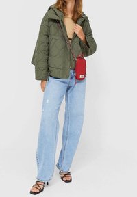 Olivgrüne gesteppte Jacke, beiges Oberteil, hellblaue Jeans mit weitem Bein und schwarze Sandalen mit Riemchen. Enthält eine kleine rote Umhängetasche.