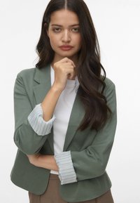 Zaļš žakets ar pielāgotu piegriezumu, kuram ir svītragaini gaiši zili piedurknes, uzvilkts virs baltas krekla ar gludu tekstūru un minimālu dekoru.