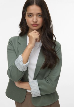 Groene blazer met een getailleerde pasvorm, voorzien van gestreepte lichtblauwe manchetten, gedragen over een witte blouse, met een gladde textuur en minimale details.