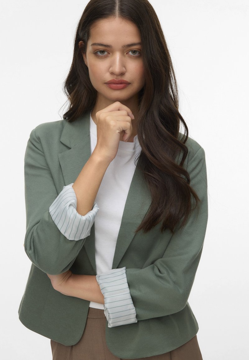 Zaļš žakets ar pielāgotu piegriezumu, kuram ir svītragaini gaiši zili piedurknes, uzvilkts virs baltas krekla ar gludu tekstūru un minimālu dekoru.