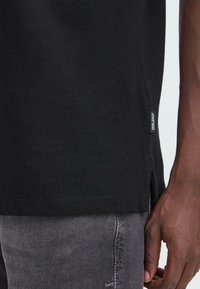 Svart bomulls T-skjorte med rund hals, sidesplitter i kanten og et lite merke nederst til venstre. Brukt sammen med grå denimjeans.