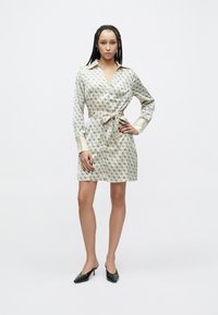 Košeľové šaty s potlačou v krémovej bielej farbe, so zavinovacím dizajnom, dlhými rukávmi a opaskom v páse. Majú saténové manžety a jemné geometrické vzory. Čierne topánky.