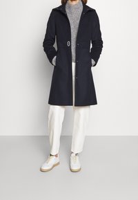 Abrigo de lana azul marino con un diseño ajustado, bolsillos frontales y herrajes plateados. Lleva encima un suéter de punto gris claro, combinado con pantalones blancos y zapatillas.