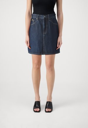 Femme portant une jupe en jean bleu foncé, un top noir sans manches et des mules noires à bouts ouverts, debout sur un fond blanc.