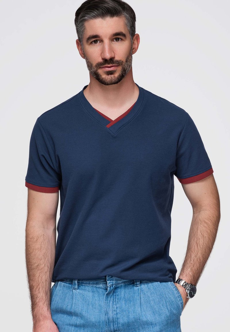 Uomo con capelli corti e scuri e barba indossa una maglietta blu navy a scollo a V con bordo rosso e jeans blu, in piedi con una mano in tasca.