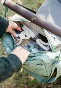 Zöld steppelt táska barna fogantyúval, amely nyitva van, és egy játékot, egy vízpalackot és egy hajtogatott szövetanyagot mutat. A belső tér csíkos anyagú.
