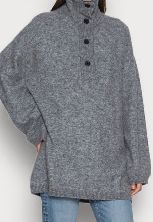 Pull en laine gris ample à col montant avec quatre boutons noirs, porté avec un jean bleu, main de la personne posée sur la hanche visible.