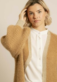 Cardigan en tricot texturé marron avec des manches larges et une garniture beige brillante, porté sur une chemise blanche à boutons.