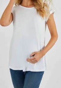 Biała koszulka damska z krótkim rękawem o luźnym kroju, gładkiej fakturze, okrągłym dekolcie i subtelnie zaokrąglonym dolnym wykończeniu, połączona z niebieskimi dżinsami.