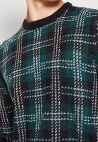 Sweter dziany z ciemnozielonym wzorem w kratę, z czarnymi i białymi akcentami, czarnym prążkowanym dekoltem i teksturowaną tkaniną.